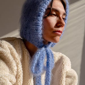May include: A person wearing a light blue fuzzy knit hat with a long tie. The hat is tied in a bow at the front of the neck. The person is wearing a white fluffy jacket.
