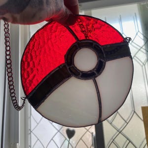 May include: A stained glass Pokeball with red and white sections, outlined in black. A chain is attached for hanging. The round design is reminiscent of a popular animated series.