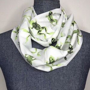 May include: White infinity scarf with a green frog print. The frogs are in various poses and have a skeletal design.