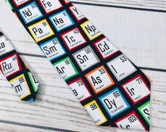 May include: A black necktie with a repeating pattern of colorful squares featuring the chemical element symbols and atomic numbers. The tie is made of a smooth fabric and has a pointed tip.