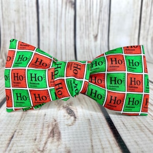 May include: A red and green bow tie with a repeating pattern of the chemical element holmium, "Ho", in a periodic table format. The bow tie is made of fabric and has a festive holiday theme.