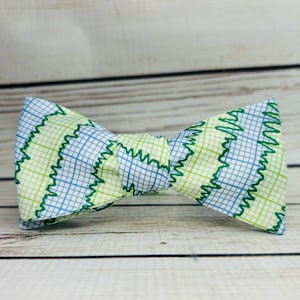 May include: A green, blue and white bow tie with a geometric pattern. The bow tie is made of patterned fabric with a grid design and green wavy lines.
