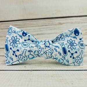 May include: A white bow tie with a blue science pattern. The pattern includes beakers, atoms, molecules, and chemical formulas.