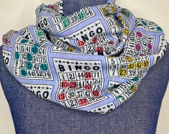 May include: A light blue infinity scarf with a repeating pattern of bingo cards. The cards have numbers and the word "Bingo" printed on them.