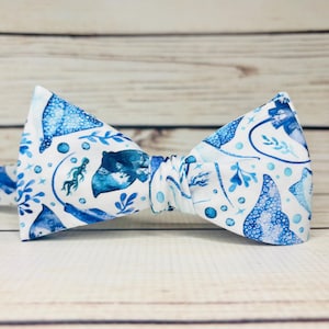 May include: A white bow tie with a blue sea life pattern, including stingrays, seaweed, and bubbles. The bow tie is made of fabric and is set against a light wood background.