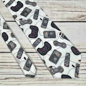 May include: A white necktie with a repeating pattern of video game controllers, handheld game consoles, and remote controls. The pattern is in shades of gray and black.