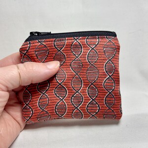 May include: A small, red zippered pouch with a repeating DNA double helix pattern in black and white. The pouch is held by a hand with a gold ring. The background is a light, neutral color.