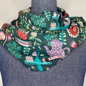 May include: A teal blue scarf with a whimsical pattern of teapots, birds, flowers, and cakes. The pattern is in shades of pink, green, purple, and white.