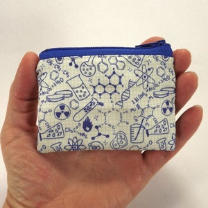 May include: A small white zippered pouch with a blue zipper. The pouch is covered in a pattern of blue science-themed illustrations, including beakers, test tubes, molecules, and chemical formulas.
