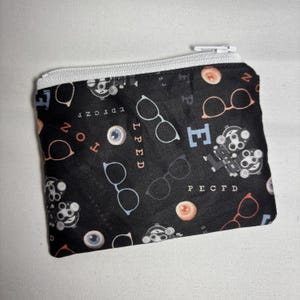 May include: A black zippered pouch with a white zipper. The pouch is covered in a pattern of eyeglasses, eyeballs, and eye chart letters in white, orange, and blue. The pouch is rectangular.