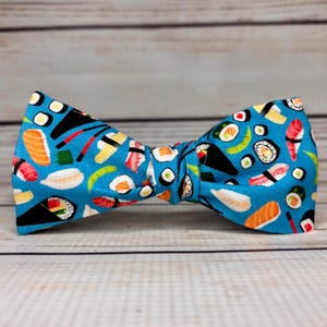 Könnte beinhalten: Eine blaue Fliege mit einem sich wiederholenden Muster aus Sushi und anderen japanischen Speisen. Die Stofffliege ist in der Mitte gebunden. Der Hintergrund ist eine helle Holzmaserung.