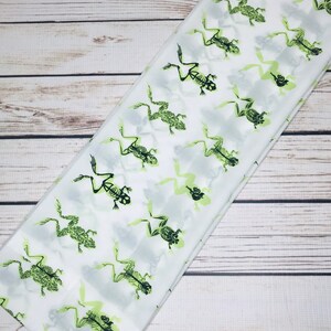May include: White fabric with a repeating pattern of green skeleton frogs. The frogs are in various poses, some are jumping, some are sitting, and some are swimming.