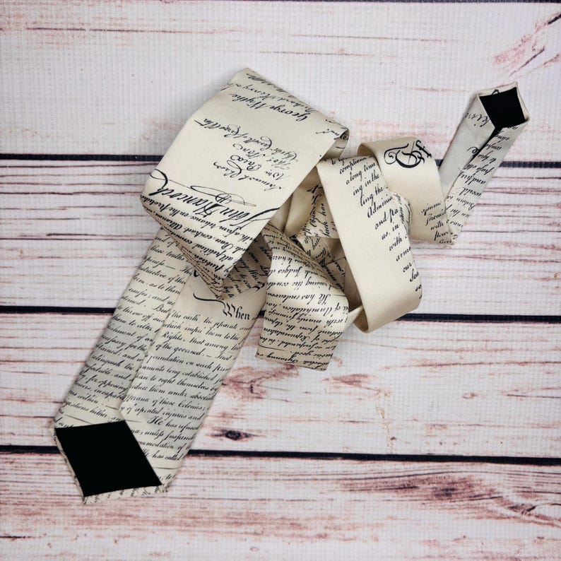 May include: A cream-colored necktie with black script resembling historical documents. The tie features a black diamond-shaped tip and is displayed on a light wood grain surface. The script includes names like John Hancock and George Wythe.