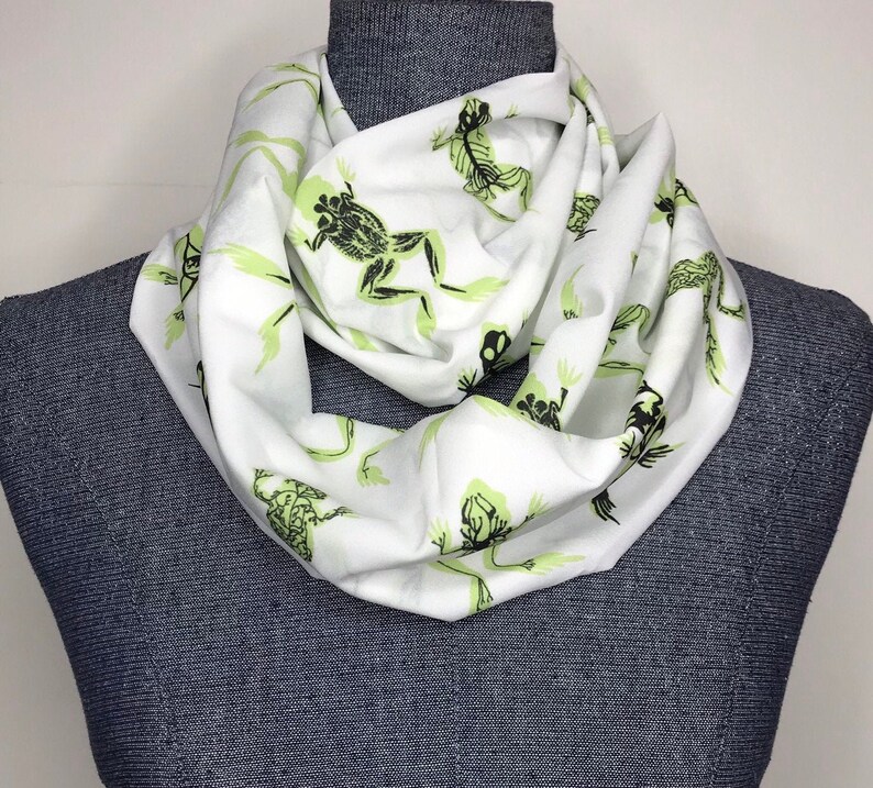 May include: White infinity scarf with a green frog print. The frogs are in various poses and have a skeletal design.