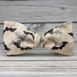 May include: A beige bow tie featuring a pattern of black and grey bats. The bow tie is knotted in the centre and made of fabric. The background is a light wood grain.