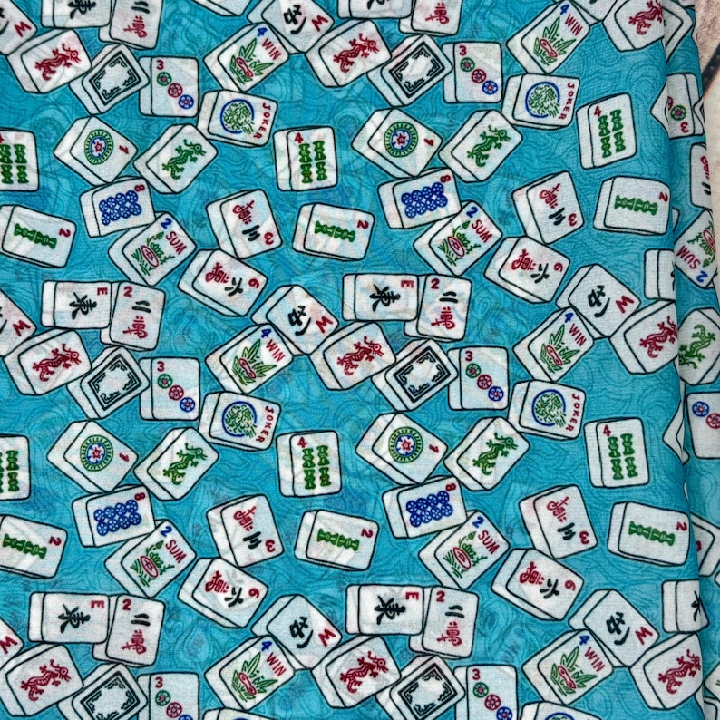 Op de afbeelding: Een turquoise blauwe stof met een herhalend patroon van witte mahjong-tegels. De tegels hebben verschillende Chinese karakters en symbolen, waaronder "WIN", "JOKER", "SUM" en "3".