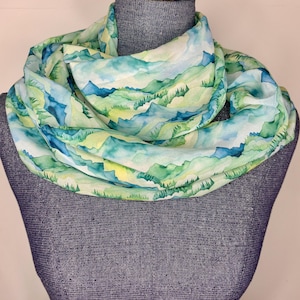 May include: A lightweight infinity scarf featuring a watercolor mountain landscape design. The scarf displays shades of blue, green, and yellow, creating a scenic pattern. The scarf is wrapped around a mannequin, showcasing its circular shape.
