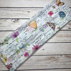 Op de afbeelding: Een wit stoffen hoofdband met een kleurrijk bloemenpatroon en wiskundige vergelijkingen en formules. Het hoofdband heeft bloemen in tinten roze, paars en geel, samen met handgeschreven vergelijkingen en formules.