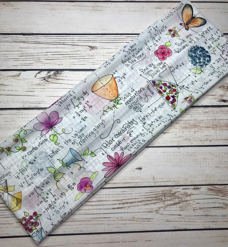 Op de afbeelding: Een wit stoffen hoofdband met een kleurrijk bloemenpatroon en wiskundige vergelijkingen en formules. Het hoofdband heeft bloemen in tinten roze, paars en geel, samen met handgeschreven vergelijkingen en formules.