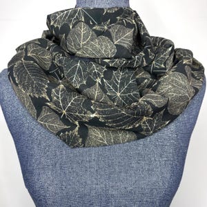 May include: A black scarf with a repeating pattern of beige leaf skeletons. The scarf is draped around a blue mannequin, showcasing the fabric's design and texture. The scarf appears to be made of a lightweight material.