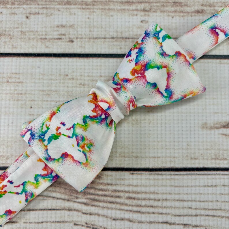 May include: A white bow tie with a rainbow world map print. The map is outlined in white and filled with rainbow colors.