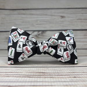 May include: A black bow tie featuring a pattern of white and red Mahjong tiles. The bow tie is made of fabric and has a classic butterfly shape. The background is a light wood grain.