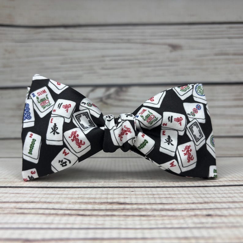 May include: A black bow tie featuring a pattern of white and red Mahjong tiles. The bow tie is made of fabric and has a classic butterfly shape. The background is a light wood grain.