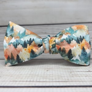 May include: A colorful bow tie featuring a watercolor mountain scene with trees, mountains, and a mix of orange, blue, and green hues. The bow tie is tied in the center and is set against a light wood grain background.