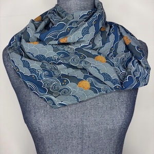 May include: A blue infinity scarf with a repeating pattern of stylized waves, moons, and stars. The scarf is made of a soft fabric and is draped over a mannequin. The design features shades of blue, white, and gold.
