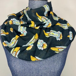 May include: Black scarf with a repeating pattern of yellow and white spaceship illustrations. The scarf is draped over a blue mannequin. The design includes small white stars on a black background.