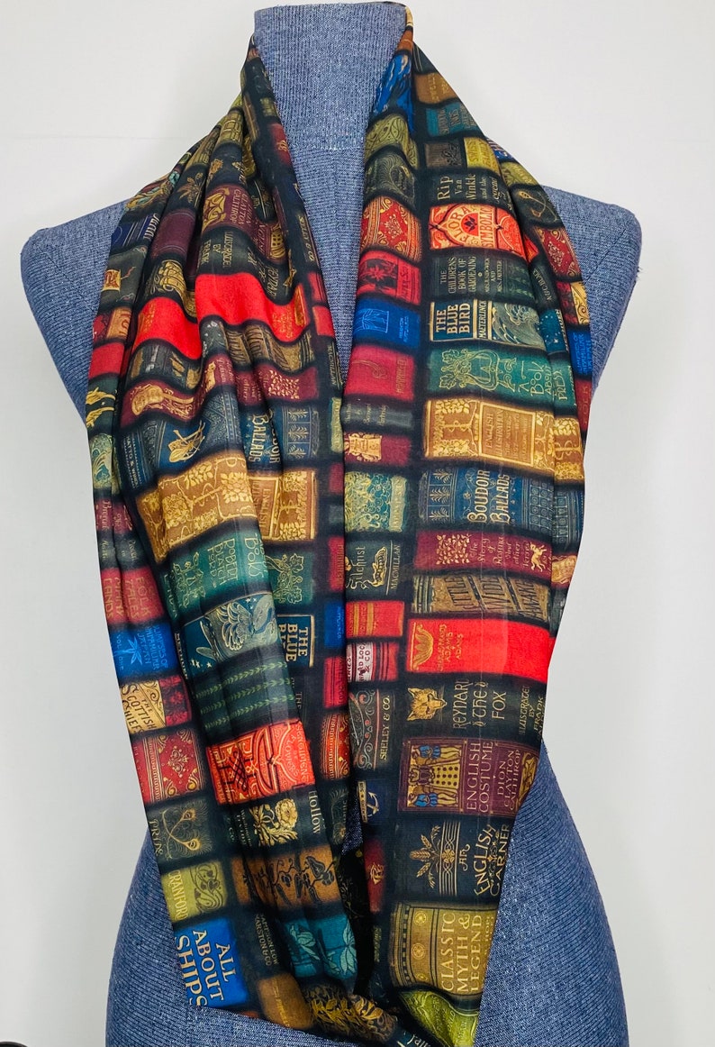 May include: A black infinity scarf with a print of colorful book spines. The scarf features a variety of book titles, including "The Blue Bird", "Boudoir Ballads", "All About Ships", and "Classic Myth & Legend".