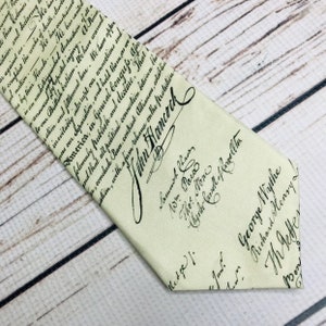 May include: Cream-colored necktie featuring handwritten text and signatures. The tie displays historical script, including the name "John Hancock" prominently. A unique accessory for history enthusiasts.