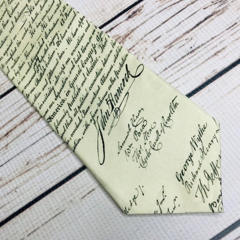May include: Cream-colored necktie featuring handwritten text and signatures. The tie displays historical script, including the name "John Hancock" prominently. A unique accessory for history enthusiasts.