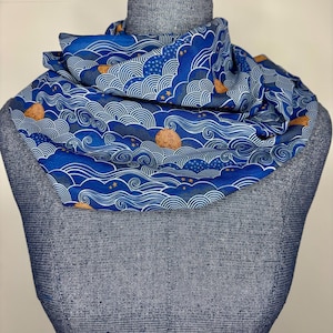 May include: A blue scarf with a repeating pattern of waves, stars, and moons. The scarf is wrapped around a mannequin's neck. The design is white and gold against a blue background.