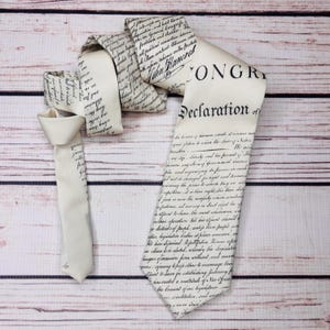 May include: Ivory-colored necktie featuring the text of the Declaration of Independence. The tie is printed with black script and has a classic pointed end. The tie is displayed on a light brown wooden surface.