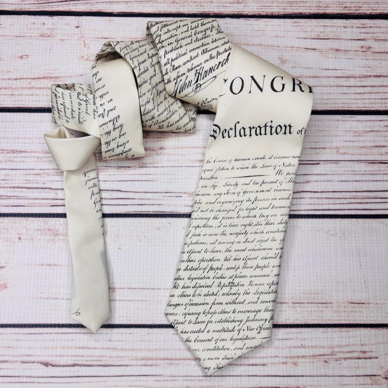 May include: Ivory-colored necktie featuring the text of the Declaration of Independence. The tie is printed with black script and has a classic pointed end. The tie is displayed on a light brown wooden surface.