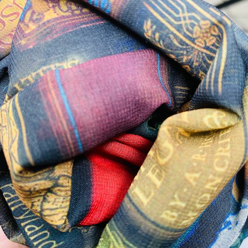 May include: A colorful scarf with a book print design. The scarf is made of a soft, silky fabric and features a variety of colors, including red, blue, gold, and black. The scarf is folded and tied in a knot.