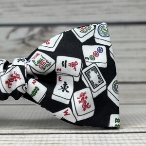 May include: A black bow tie featuring a pattern of white Mahjong tiles with red, green, and blue symbols. The bow tie is made of fabric and is tied in a classic bow shape. The background is a light wood.