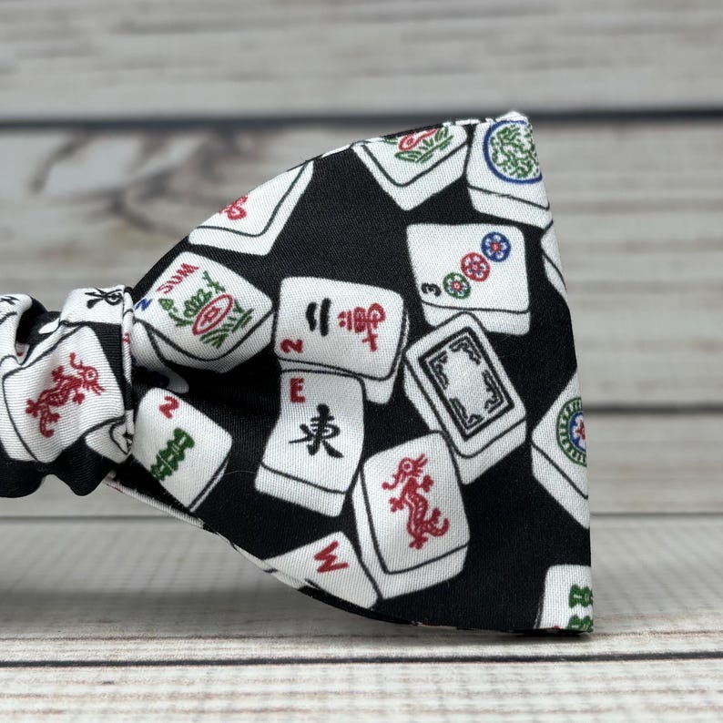May include: A black bow tie featuring a pattern of white Mahjong tiles with red, green, and blue symbols. The bow tie is made of fabric and is tied in a classic bow shape. The background is a light wood.