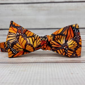 May include: A bow tie with a repeating pattern of monarch butterflies. The butterflies are in shades of orange, black, and yellow. The bow tie is made of fabric and is tied in the centre. The background is light wood.