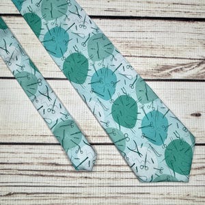 May include: A light blue tie with a pattern of green circles and surgical instruments. The tie is made of a silky material and has a pointed tip.