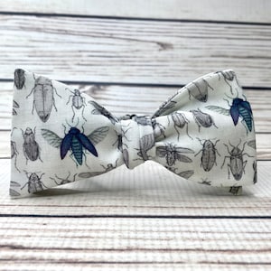 May include: A white bow tie featuring a pattern of black and blue insects. The bow tie is crafted from a soft, textured fabric.