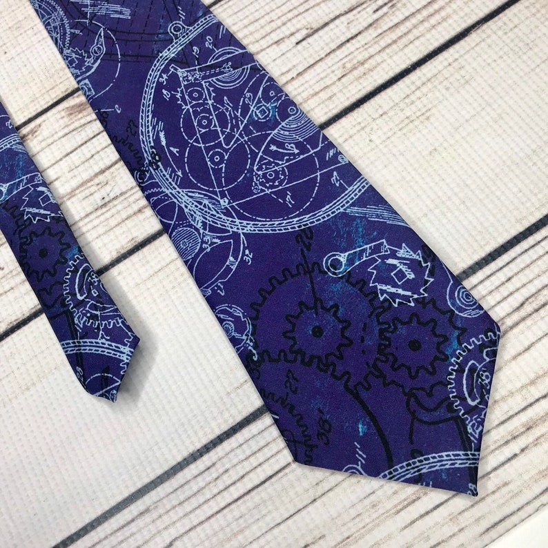 Tie front and end lay parallel angled into the corner of the frame. the tie design is a dark blue background with lighter shades of blue making a parchment-like look, on yop of that are white and black gears, cogs, and other machinery diagrams.