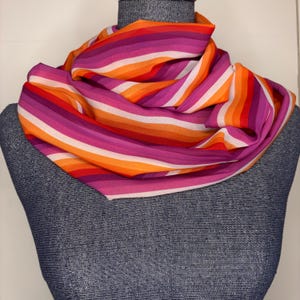 May include: A colorful scarf with horizontal stripes in shades of orange, red, pink, purple, and white. The scarf is draped over a mannequin, showcasing its flowing fabric and vibrant color palette. The scarf's design is eye-catching and suitable for various occasions.