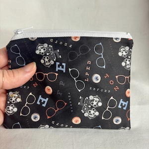 May include: Black zippered pouch with a pattern of eyeglasses, eye charts, and eyeballs. The design includes eyeglasses in white and orange, eye charts, and the letters E, F, and P. The zipper is silver.