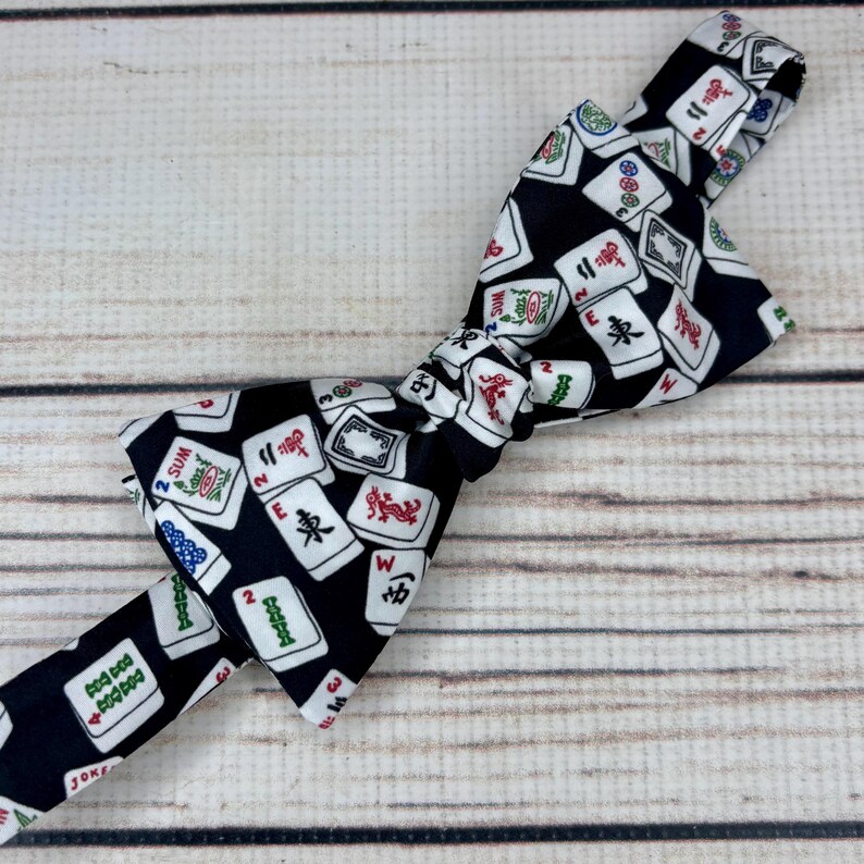 May include: A black bow tie featuring a pattern of white Mahjong tiles with red, green, and blue accents. The bow tie is tied in the center and lies on a light brown wooden surface.