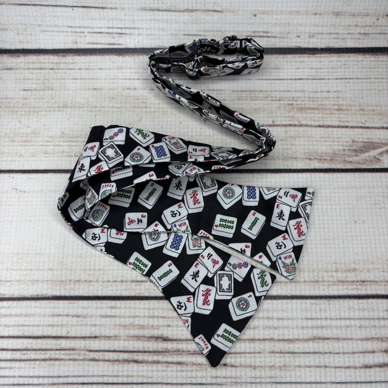 May include: A black bandana and matching headband featuring a pattern of white and red Mahjong tiles. The bandana is triangular, and the headband is elasticated. Both are on a wooden surface.