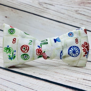 Pale wood background. Bowtie is tied and standing up, it has a very pale, creamy green background, and the tiles are outlined in a darker green. The Mahjong tiles have red and green numbers, wth designs that are red green, and blue.