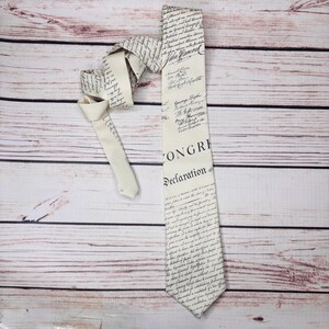 May include: Cream-colored necktie featuring a printed design of historical documents, including the text "CONGRESS Declaration". The tie is knotted and laid on a wooden surface. The design includes signatures and script.