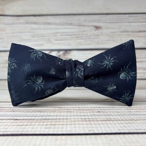 May include: A dark navy bow tie with a repeating pattern of grey spiders. The fabric bow tie is tied in the centre. The background is a light-coloured wooden surface.